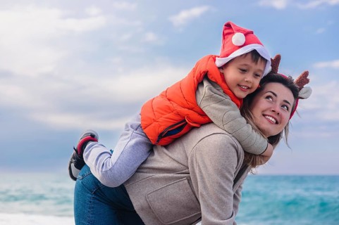 A young Caucasian woman with brown hair carries her smiling son on her back at the beach during