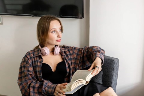 Young Caucasian woman with long brown hair, wearing a black top and plaid shirt, sitting on a gray