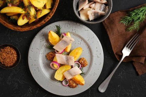 Plate with potato wedges alongside slices of herring. Small bowls hold mustard and fresh herbs.