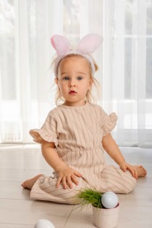 A toddler girl sits on the floor in a bright room. She wears bunny ears and smiles while surrounded