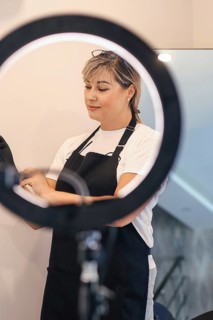 Woman adjusting gloves under a ring light, wearing an apron, appears focused, professional
