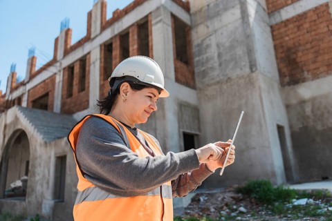 A young Hispanic woman in a hard hat and orange vest uses a tablet at a construction site. The