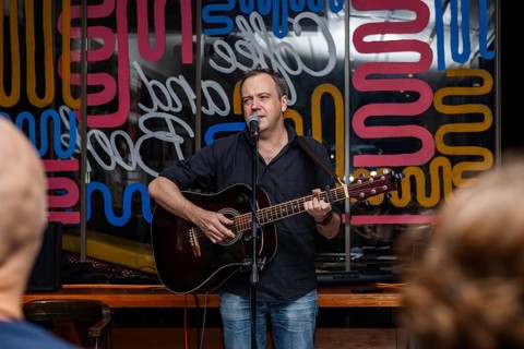 A man with short light brown hair sings and plays guitar on stage. Colorful abstract background.