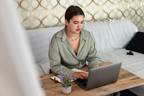 A young Hispanic woman with long brown hair works on a laptop at a wooden table in a cozy living
