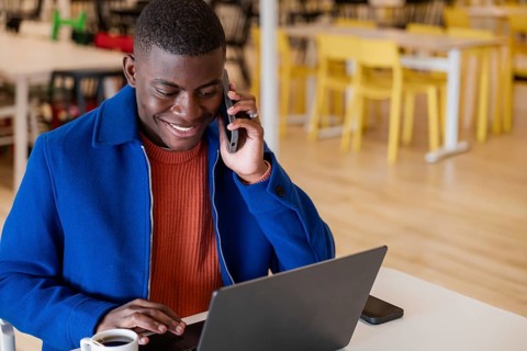 A young African man with short hair is sitting at a table in a cafe. He is smiling while talking on