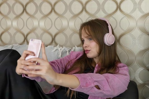 Young Caucasian woman with long brown hair wearing pink headphones, sitting on a couch and using a