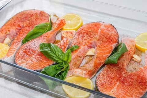 Fresh salmon fillets arranged in a glass dish with lemon slices and basil leaves. The fish is raw