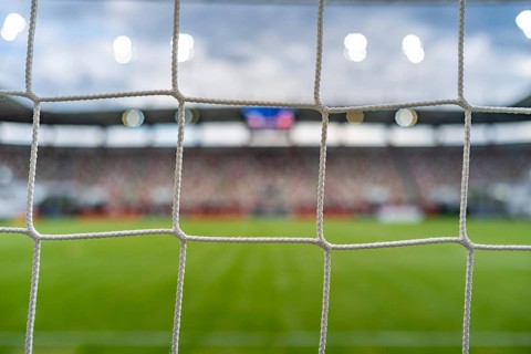Soccer goal net in the foreground with a blurred football field, stadium and spectators in the
