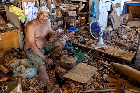 Man sitting in chaotic workshop holding a carved fish sculpture, craftsman leg carving masks in