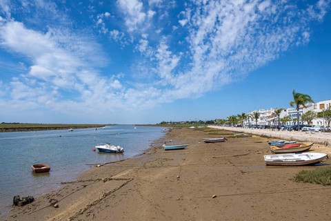 Boats lie on the sandy shore of a lagoon with blue skies and scattered clouds, flanked by white