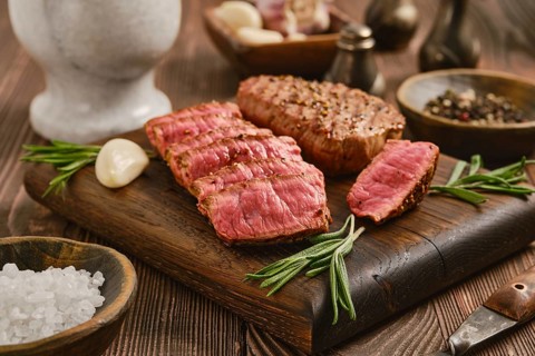 Beef steak is sliced and displayed on wooden board. Whole garlic cloves and bowls of spices are