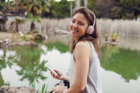A young Hispanic woman with long brown hair smiles while wearing headphones. She holds a smartphone