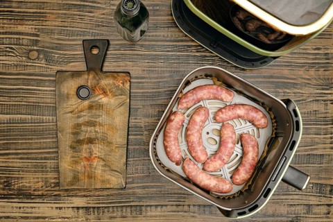 Banger sausages in air fryer basket on kitchen counter. Wooden cutting board is placed next to air