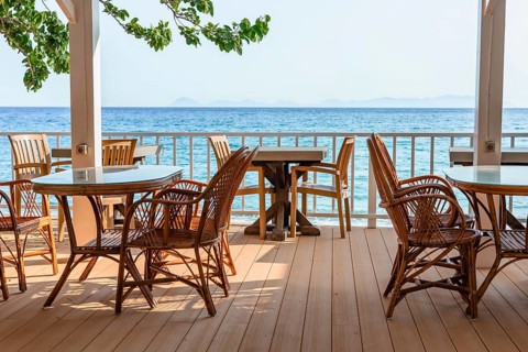 Rattan chairs and tables set on a seaside terrace overlooking clear blue water on a bright and