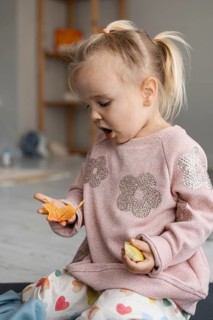 A young Caucasian girl with blonde hair in pigtails sits on the floor, holding orange leaves. She