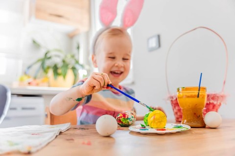 A child wearing bunny ears sits at a table decorated for Easter. Colorful eggs are placed on the