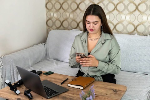 A young Caucasian woman with long brown hair sits at a wooden table, using a smartphone. A laptop,