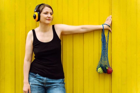 Cheerful woman with fresh veggies in mesh tote, urban style on yellow, Woman wearing headphones