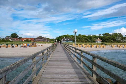 A long wooden walkway leads across the sea with a view of beach houses and beach chairs under a