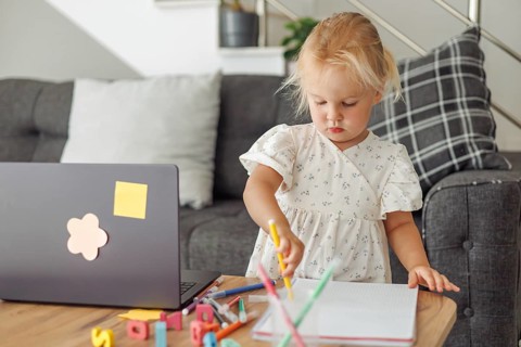 Young blond European girl drawing with colored pencils at home near laptop and sofa in living room