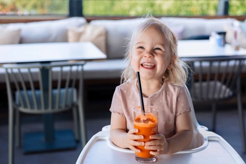 Happy smiling girl toddler drinking carrot juice with a straw in cafe