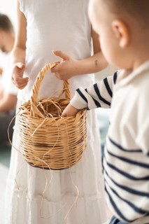 A young boy with short hair reaches into a woven basket held by a girl in a white dress. The scene