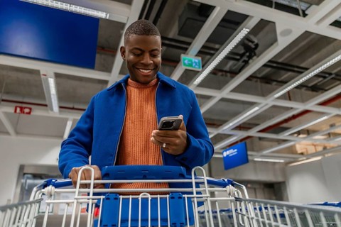 A young African man smiles while using a smartphone in a shopping cart. The scene is set in a