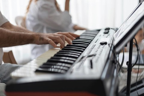 A close-up of a man's hand playing a keyboard in a cozy home setting. A woman is seated nearby,