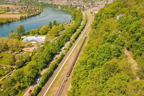 Aerial view of a railway line next to a river and a city, surrounded by green hills, construction