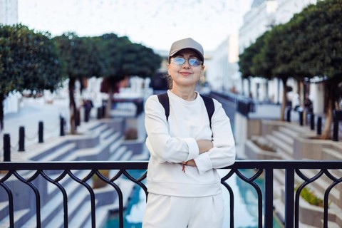 A woman wearing a cap and glasses stands with her arms crossed near a beautifully designed water