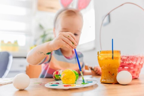 Toddler girl in dress wearing bunny ears painting Easter egg with brush, basket for eggs is near