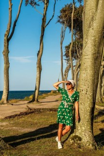 A woman (54) in a green tennis dress and white trainers is leaning casually against a tree, gazing
