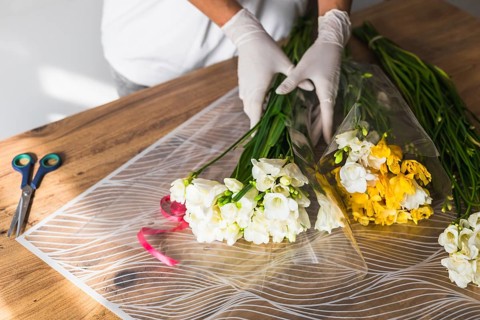 A florist in gloves carefully wraps a bouquet of white flowers with yellow accents on a wooden
