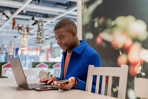 Young African man using a laptop and smartphone in a modern cafe. Bright interior with decorative