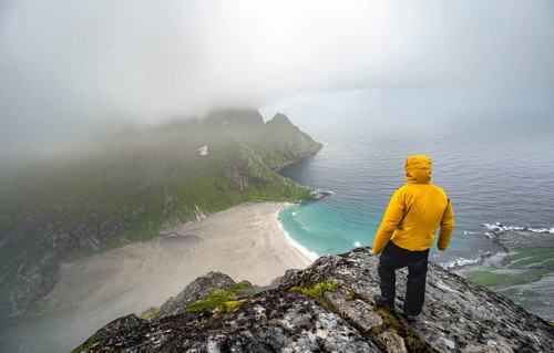 Hiker on Helvetestinden summit, view of Bunesstranden sandy beach and blue sea, dramatic cloudy