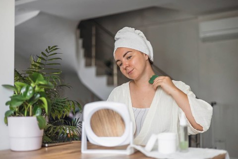 A young Caucasian woman with a towel on her head performs gua sha face massage in a bright room.