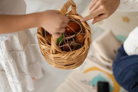A childs hand reaches for a woven basket filled with colorful eggs. The scene captures a warm,