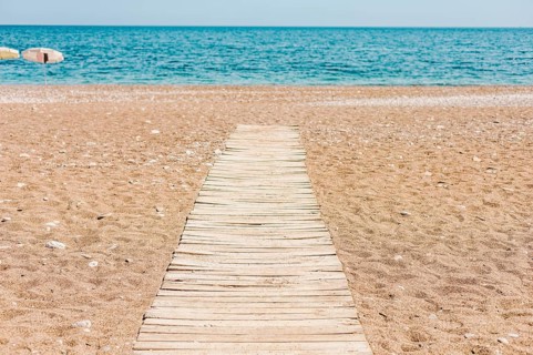 A wooden pathway leads to a calm beach with golden sand and turquoise water. The sky is clear,