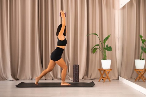 A woman stands on a yoga mat holding dumbbells, facing a mirror in an exercise studio