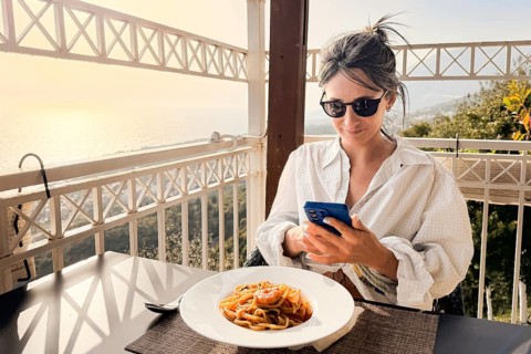 Woman taking photo eating shrimp spaghetti on scenic Mediterranean restaurant terrace. Golden hour