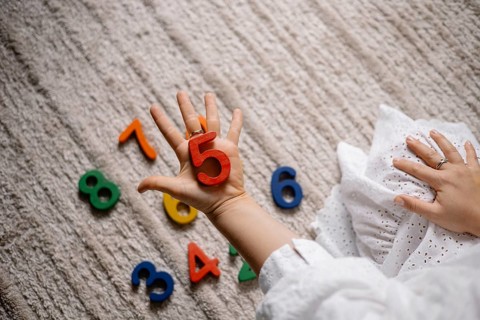 Child holding a red number five with other colorful numbers scattered on the floor