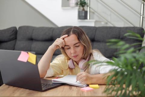 Young blond European woman with headphones sitting at wooden table with laptop and notebook,