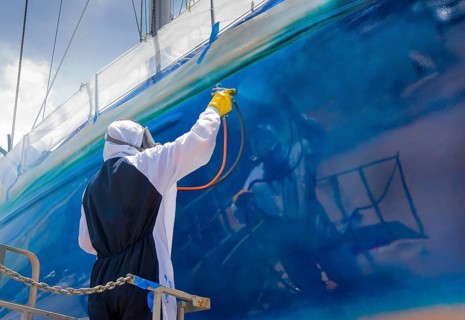 Worker spray paints the hull of a sailing yacht during professional marine maintenance, wearing