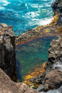 Natural rock pool filled with clear turquoise water, surrounded by dark volcanic rock formations,