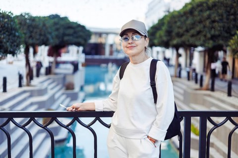 Middle-aged woman stands casually by a railing, smiling while overlooking a serene water feature in