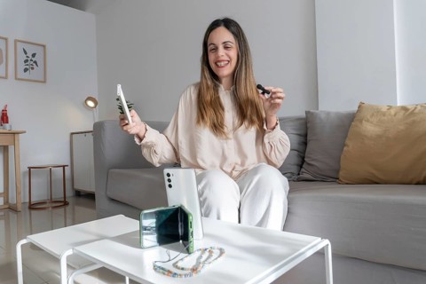 Cheerful woman sitting on couch holding up smartphone and lavalier microphone, phone with beaded