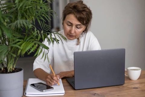 Serious pretty mature woman in white outfit studying at laptop at home, writing notes in notebook,