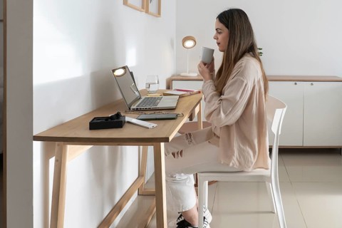 Mid adult woman sitting at desk taking a coffee break, holding mug and looking at laptop screen,