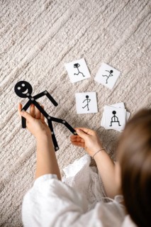 Hands holding a stick figure toy with cards featuring poses on a textured floor