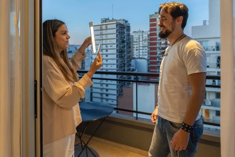 Woman holding LED light panel and smartphone up to smiling man's face on balcony at dusk, city